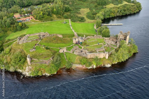 Aerial view of the historic ruins of Urquhart Castle on the banks of Loch Ness with green grounds and visitors walking along paths in Drumnadrochit, Scotland, United Kingdom.