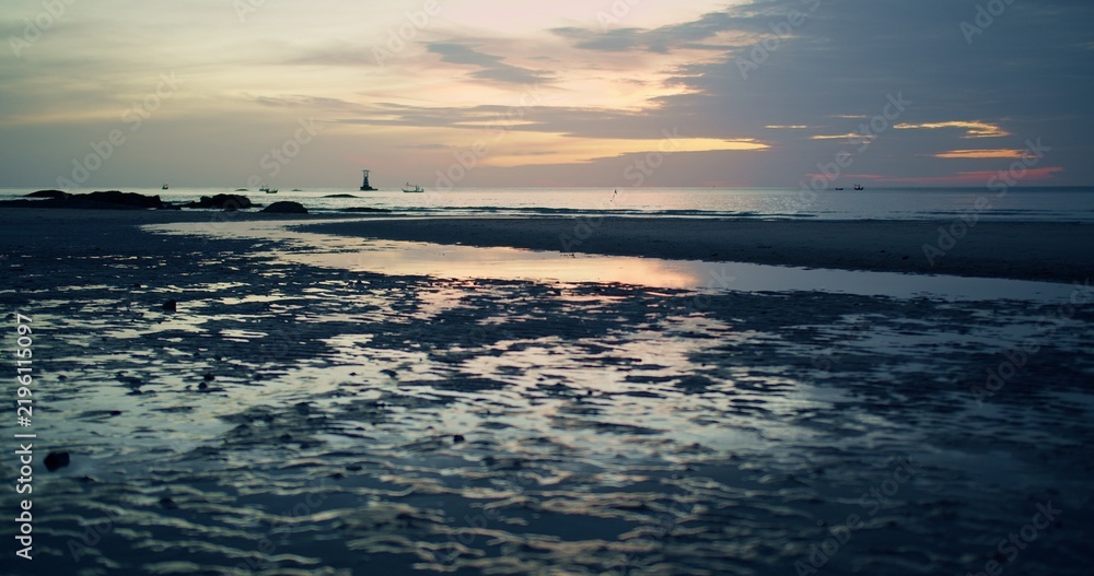 Fototapeta premium A serene coastal landscape at low tide reflects the beautiful twilight sky in tidal pools, with distant fishing boats on the calm ocean creating a peaceful travel scene.