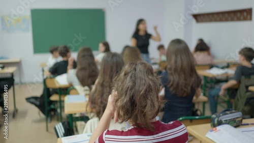 Students sitting at desks in a classroom with a teacher standing in front of a green chalkboard