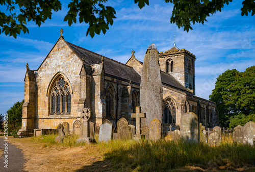 Foto Historic All Saints’ Church in Rudston, East Yorkshire, with the remarkable prehistoric Rudston Monolith rising among old gravestones in the churchyard on a sunny summer day
