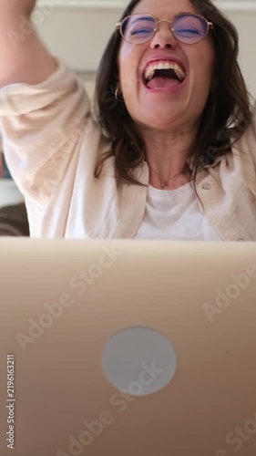 Happy professional woman looking at her laptop screen, celebrating a successful business deal, raising her arms in victory, and screaming with excitement in her modern home office