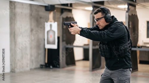 Adult man practicing pistol marksmanship at indoor shooting range wearing tactical gear and aiming at paper target