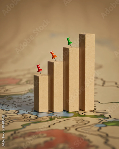 Wooden bar chart showing upward growth trend with colorful pushpins marking milestones arranged sequentially upon an old vintage map surface representing business strategy analysis and financial