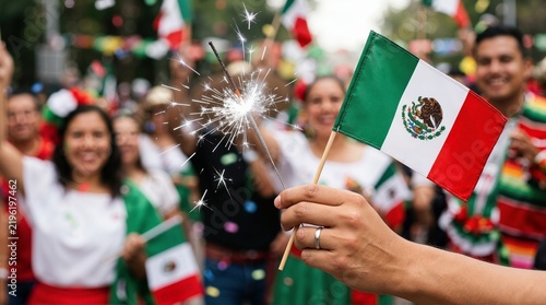 Hand holding small Mexican flag and burning sparkler during outdoor fiesta. Mexican Independence Day street parade