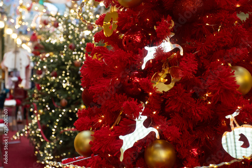 vibrant red christmas tree covered in gold baubles and reindeer ornaments,illuminated by warm fairy lights,with sparkling green trees blurred behind in a festive shopping arcade