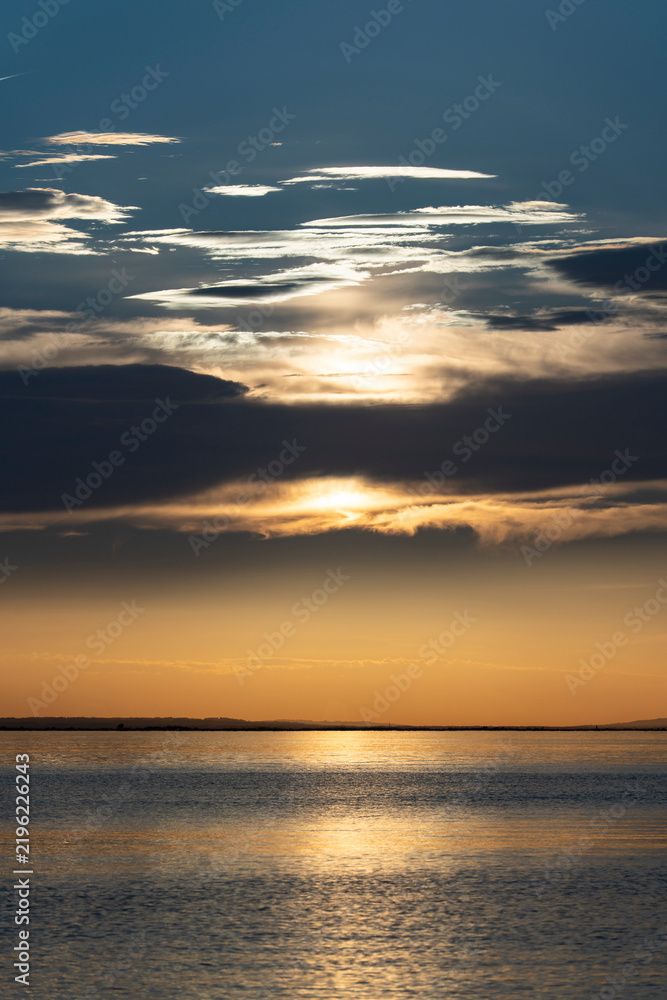 Fototapeta premium atmospheric sunset view over Lake Constance (Bodensee) taken from the Austrian shore in Bregenz, featuring golden sunlight breaking through dramatic cloud layers and reflecting gracefully across the c