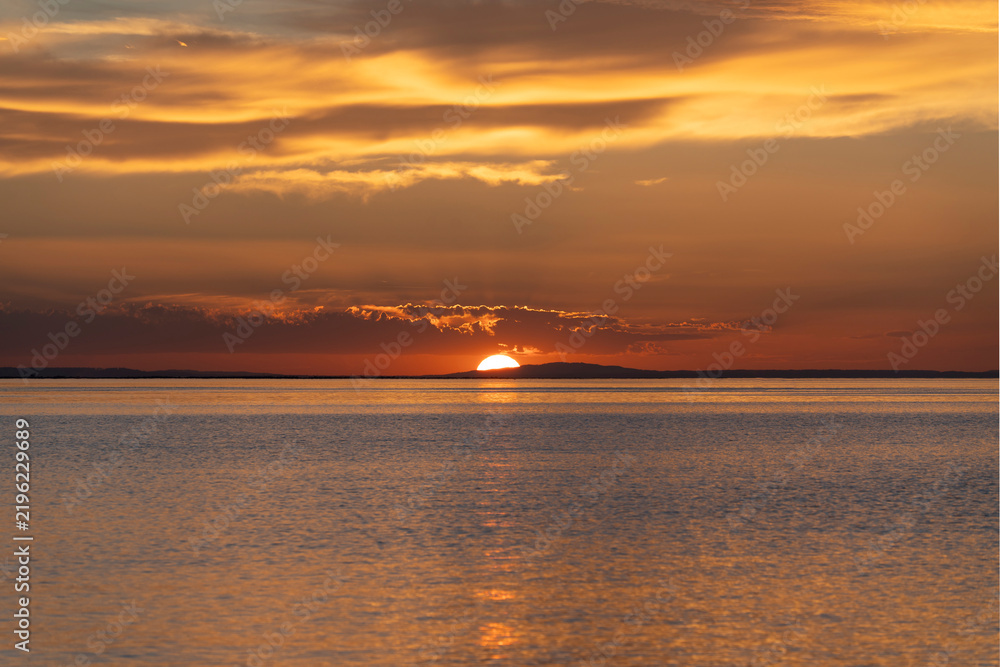 Fototapeta premium sunset over Lake Constance captured from the shoreline of Bregenz, Austria, with the setting sun dipping behind distant hills and casting a brilliant golden glow over the calm water