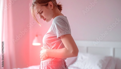 Female person suffering from sharp abdominal cramps and lower backache inside a sunlit minimalist bedroom.