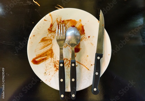 Empty dirty white plate with fork, spoon, knife and food stain after meal on black background, top view