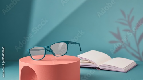 Two pairs of glasses resting on a pink cylindrical podium next to an open book on a blue background.