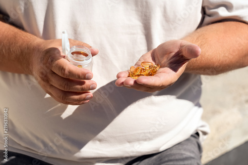 Overweight man holding handful of golden omega-3 fish oil gel capsules and open bottle, daily supplement routine, obesity healthcare, weight management, and metabolic support