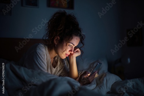 Young woman lying in bed at night looking at smartphone screen, face illuminated by screen light