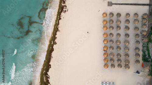 Aerial view of Playa del Carmen beach with rows of straw umbrellas on white sand, turquoise water, and seaweed along the shoreline in Playa del Carmen, Quintana Roo, Mexico.