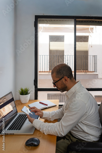 Young man checking smartphone beside laptop at desk
