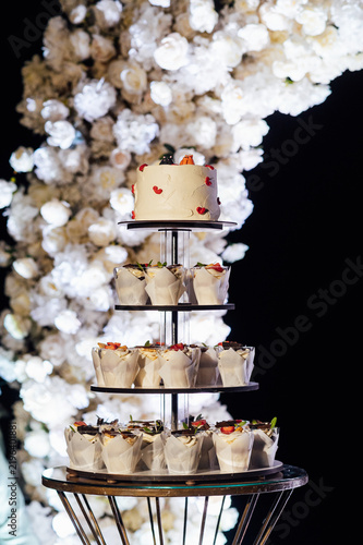 Tiered wedding cake and cupcake tower stand in front of illuminated floral arch at night.