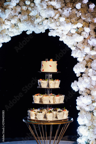 Tiered wedding cake and cupcake tower stand in front of illuminated floral arch at night.
