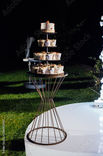 Tiered wedding cake and cupcake tower stand in front of illuminated floral arch at night.