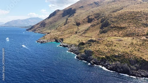Coastal landscape featuring rocky shoreline and clear blue waters, with rolling hills and distant mountains under a bright sky, showcasing the natural beauty of the Mediterranean region