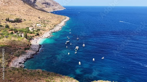 Aerial view of a serene sea coast with a beach, yachts anchored in clear blue waters, and a rugged landscape featuring hills and vegetation under bright sunlight. Sea resort at warm summer day