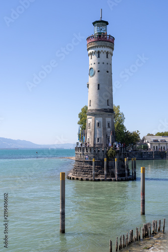 a scenic view of the historic harbor entrance to Lindau on Lake Constance in Germany, featuring the famous New Lighthouse beneath a bright blue sky and turquoise waters