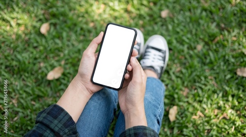 High angle view of hands holding a black smartphone with a blank white screen in a park setting.