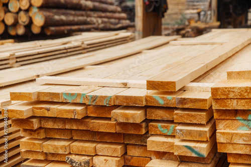 Stacked wooden planks in a lumber yard with logs and timber in the background, showcasing various sizes and textures of untreated wood materials