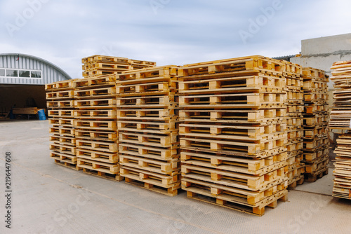 Stacked wooden pallets arranged neatly in an industrial yard with a warehouse structure visible in the background under a cloudy sky