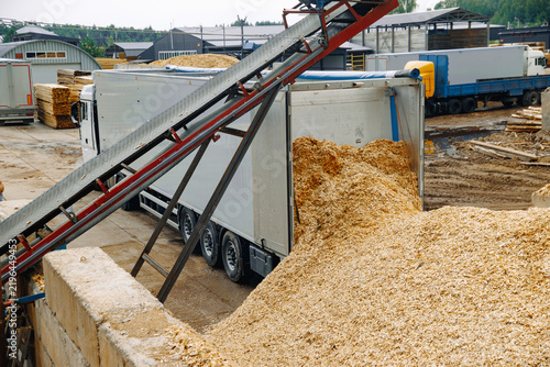 Truck unloading wood chips at a lumber yard with conveyor belt system and stacks of lumber in the background under a cloudy sky