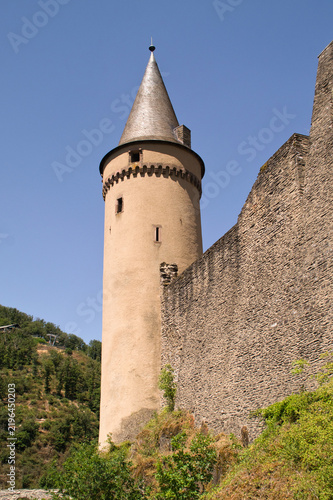 Medieval castle tower rises beside a massive stone fortress wall under a clear blue sky. The architectural landmark showcases European heritage