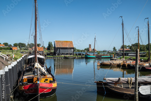 Historic wooden sailing ships and flat-bottomed vessels moored in the harbor with traditional smokehouse chimneys in the background at the open-air museum in Enkhuizen.