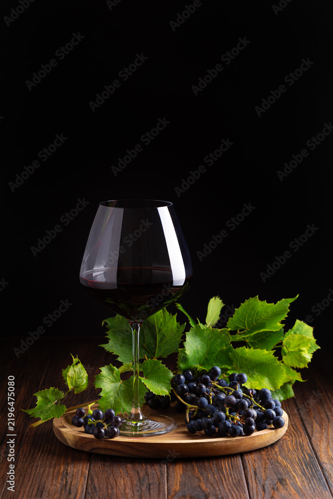 Obraz premium Elegant glass of red wine with fresh dark grapes and green leaves on a rustic wooden surface