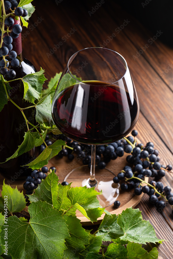 Obraz premium Elegant glass of red wine and bottle surrounded by fresh grapes and leaves on a rustic wooden table