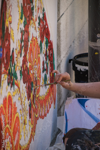Quadro su tela Extreme close-up shot focused on an artist's hand holding a small paintbrush, precisely applying red paint to a flower detail on a larger, textured mural wall