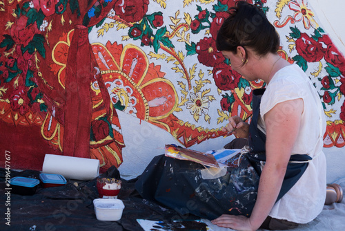 Profile view of a young female painter sitting outdoors, carefully painting intricate floral details on a large mural project.