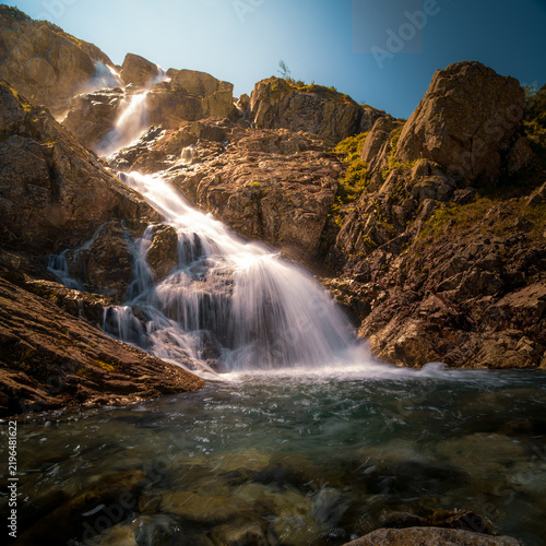 A breathtaking mountain waterfall cascading over rugged rocks amidst pristine alpine scenery. Surrounded by lush vegetation and towering peaks, the flowing water creates a peaceful and refreshing atmo