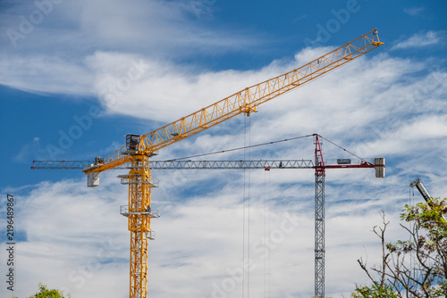 Fotografie A bright yellow construction tower crane extends diagonally against a vibrant blue sky with wispy clouds alongside a second crane mast