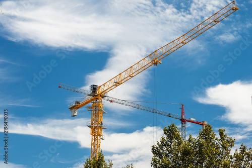 Cuadro en lienzo A bright yellow tower crane with an extended jib rises above green treetops under a vibrant blue sky dotted with fluffy clouds