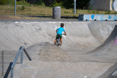Fotografie A young boy on a bicycle rides up an inclined ramp beside metal grind rails at a concrete outdoor skatepark