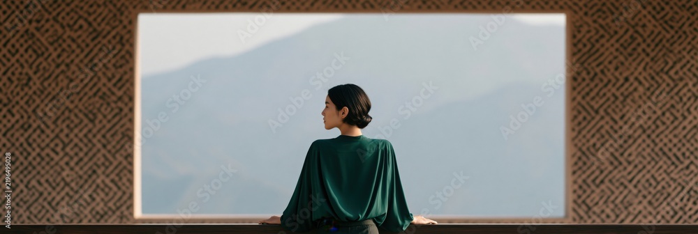 Obraz premium Woman standing at large framed window overlooking misty mountain view for serene minimalist travel wallpaper and meditation background