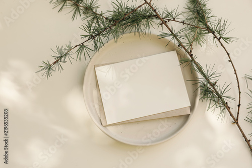 Neutral Christmas stationery mockup with blank horizontal greeting card invitation, ceramic plate. Larch tree branche, white table in sunlight. Neutral Scandinavian holiday background, winter flat lay