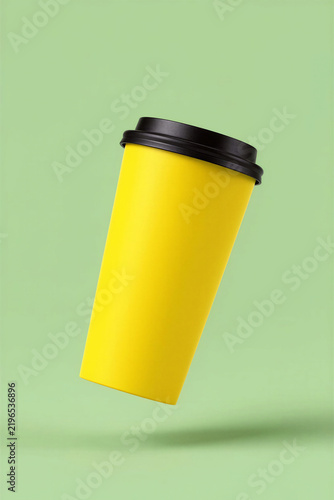 Vivid golden yellow takeaway coffee cup with black snap-on lid floating at an angle against a pastel green background, minimalist beverage packaging mockup, copy space
