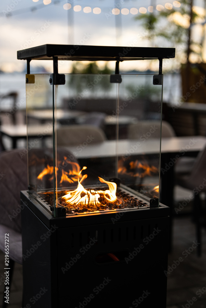 Fototapeta premium Modern outdoor gas fireplace with flames inside glass box for guests comfort at trendy restaurant patio, stylish gas heater with fire behind glass at cafe open-air terrace, cozy lounge settings.