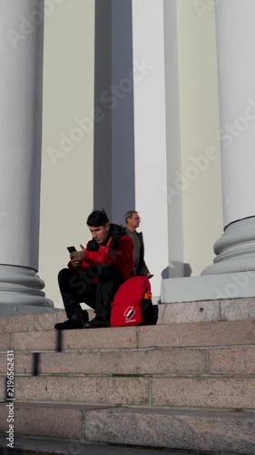 Man in Red Jacket Sitting in Front of a Building Looking at His Phone
