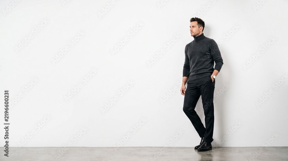 Fototapeta premium Professional man in dark sweater and jeans leaning casually against plain wall with clean copy space