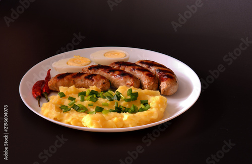 Bild auf Leinwand Grilled sausages with mashed potatoes and a boiled egg, with sliced ??green onions and chili pepper in a round plate against a black background