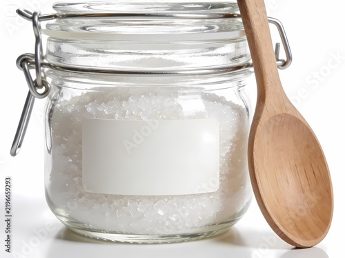 Airtight glass jar filled with white sugar or sea salt crystals with a blank label and wooden spoon on a white background