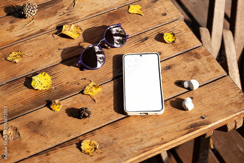 Smartphone with blank screen, sunglasses and wireless earbuds on wooden table with yellow autumn leaves.