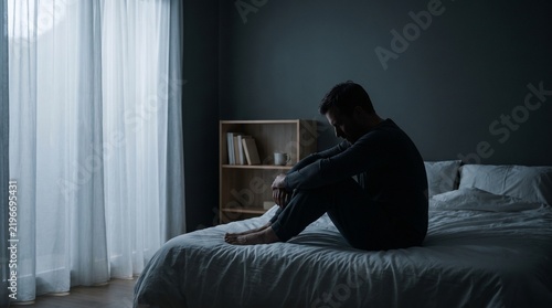 Loneliness and Emotional Wellbeing Concept with Man Sitting Alone on Bed in Quiet Dark Bedroom Interior