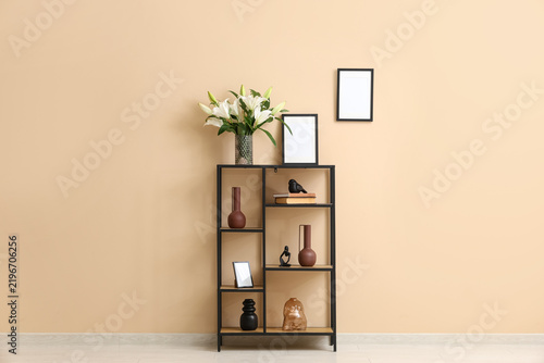 Shelving unit with vase of lily flowers and photo frames near beige wall in room