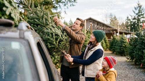 A young family securing a Christmas tree tied to car roof at an outdoor winter farm